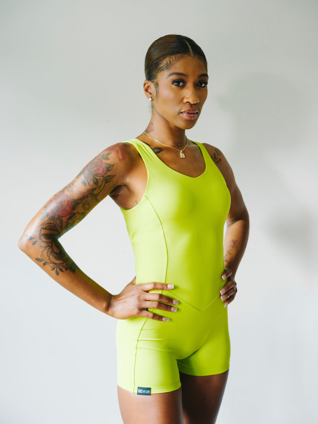 A woman modeling a bright neon yellow-green athletic one-piece bodysuit with matching shorts against a light gray studio background.