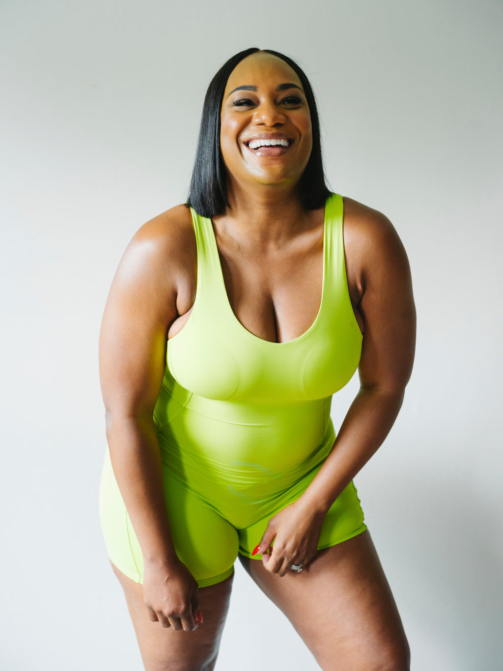 A smiling woman wearing a bright neon yellow-green one-piece bodysuit with matching shorts, photographed against a plain light gray studio background.