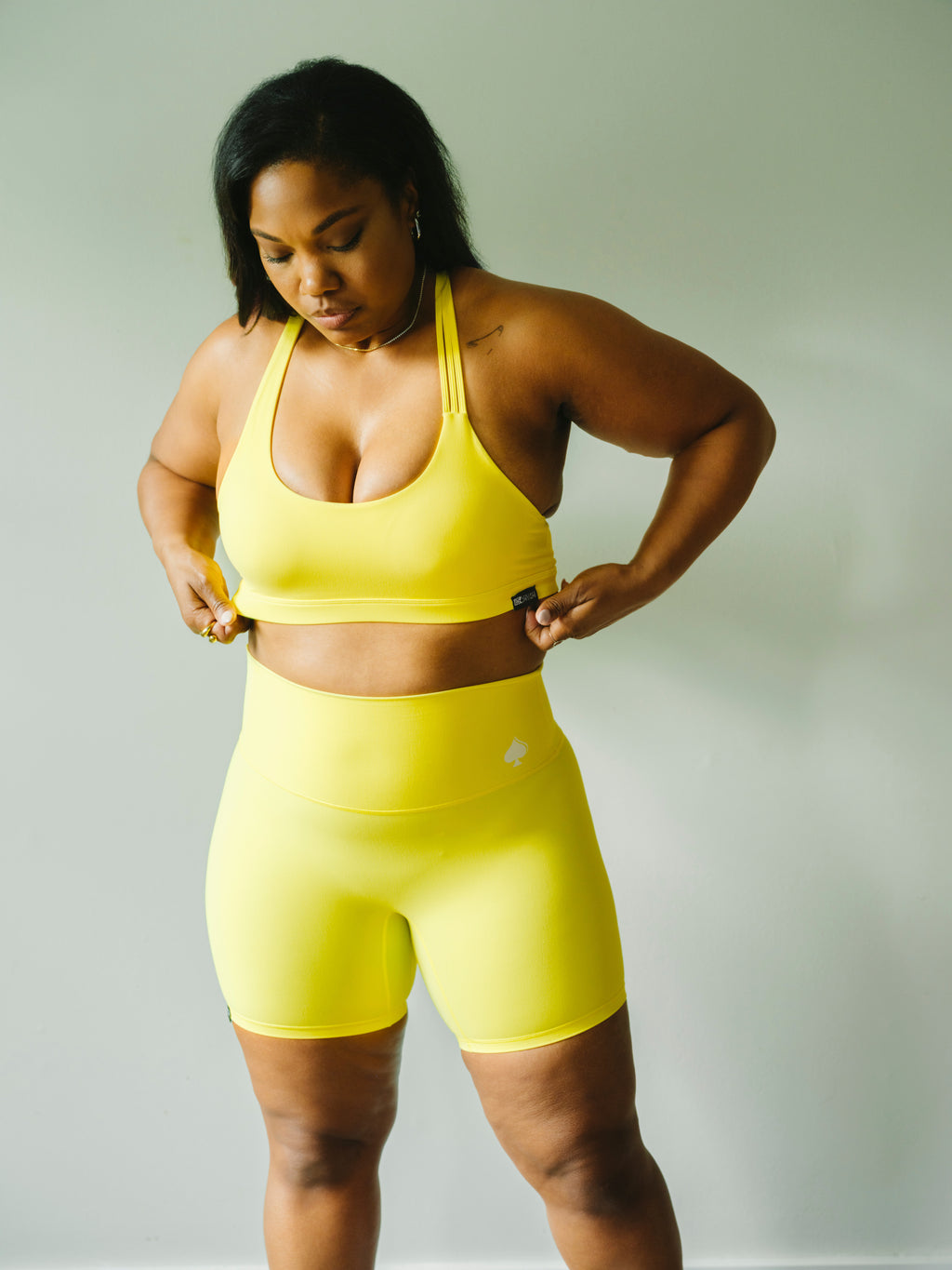 A woman modeling a matching neon yellow activewear set consisting of a sports bra and high-waisted biker shorts, standing against a plain light gray background.