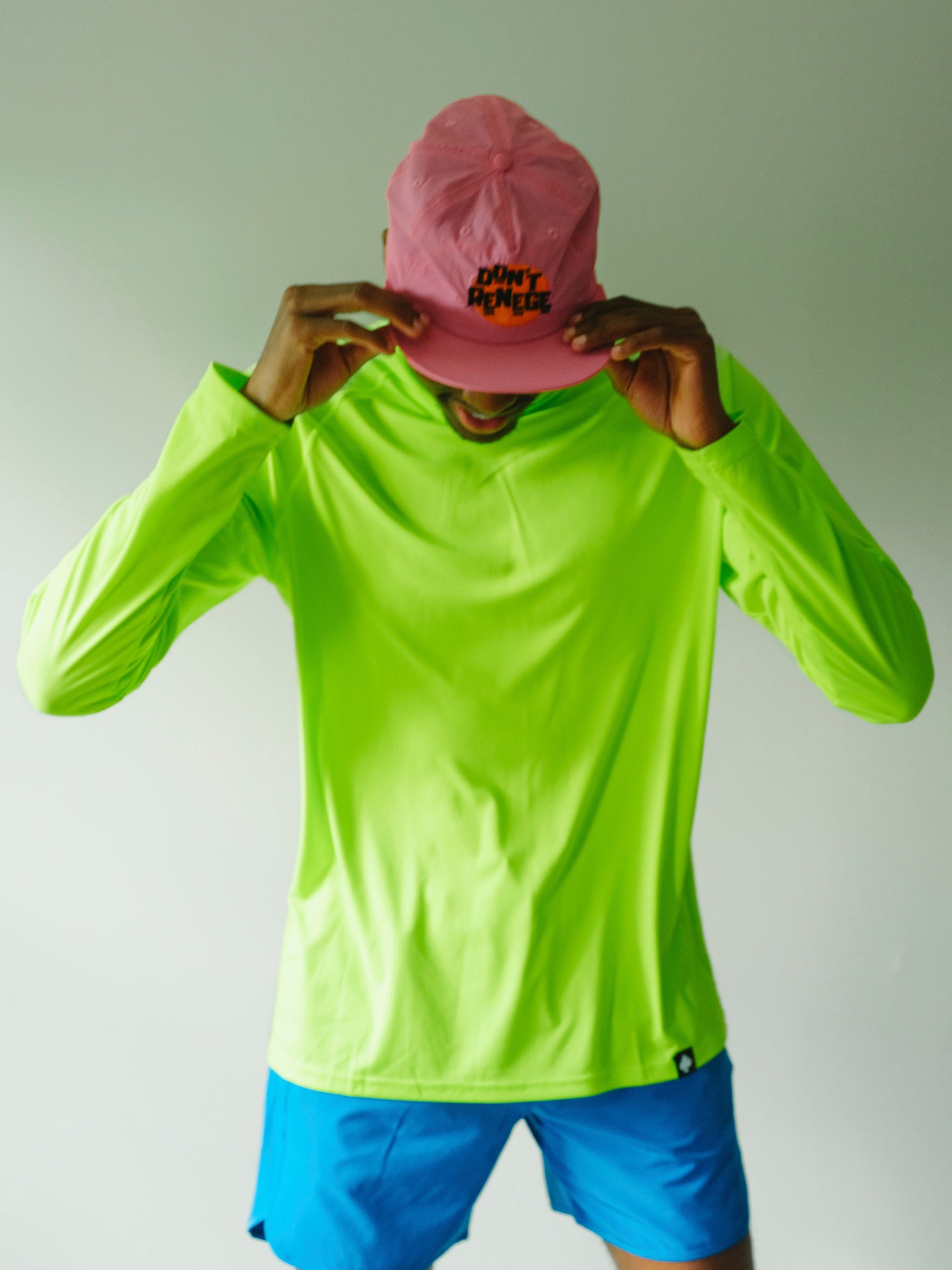 A person wearing a bright neon yellow-green long-sleeve athletic shirt, blue shorts, and a pink baseball cap with 'SUNY DUNKIN' printed on it, holding the cap in front of their face against a plain light gray background.