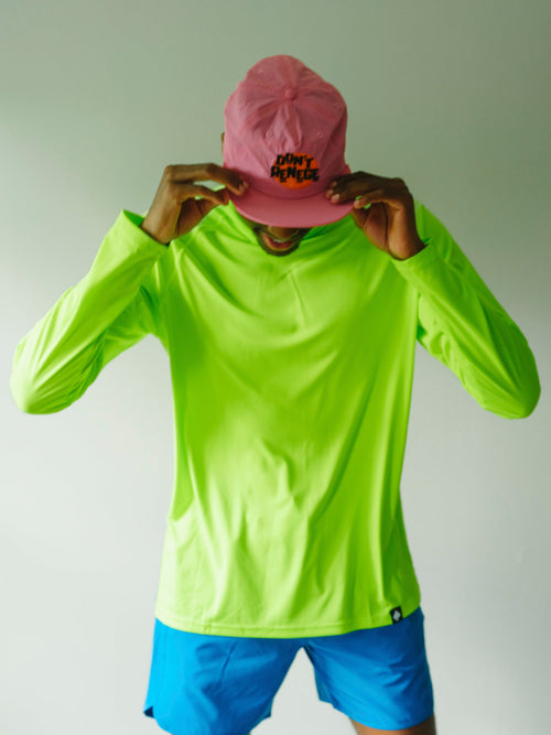 A person wearing a bright neon yellow-green long-sleeve athletic shirt, blue shorts, and a pink baseball cap with 'SUNY DUNKIN' printed on it, holding the cap in front of their face against a plain light gray background.