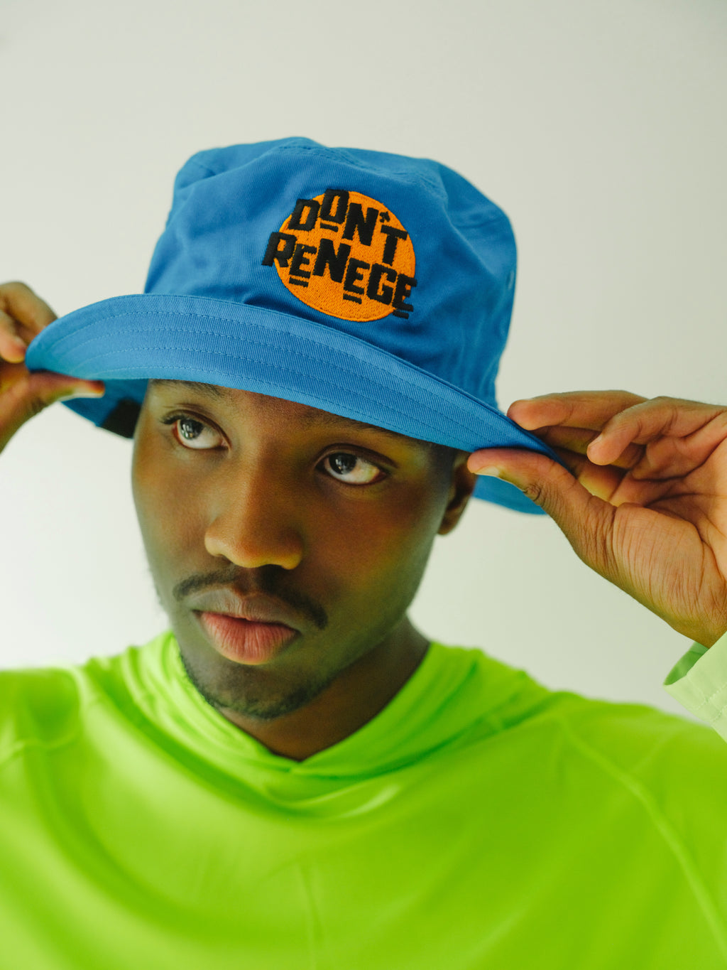 A man wearing a bright blue bucket hat with a circular orange and black 'DON'T RENEGE' graphic on the front, holding the brim with both hands against a plain light background.