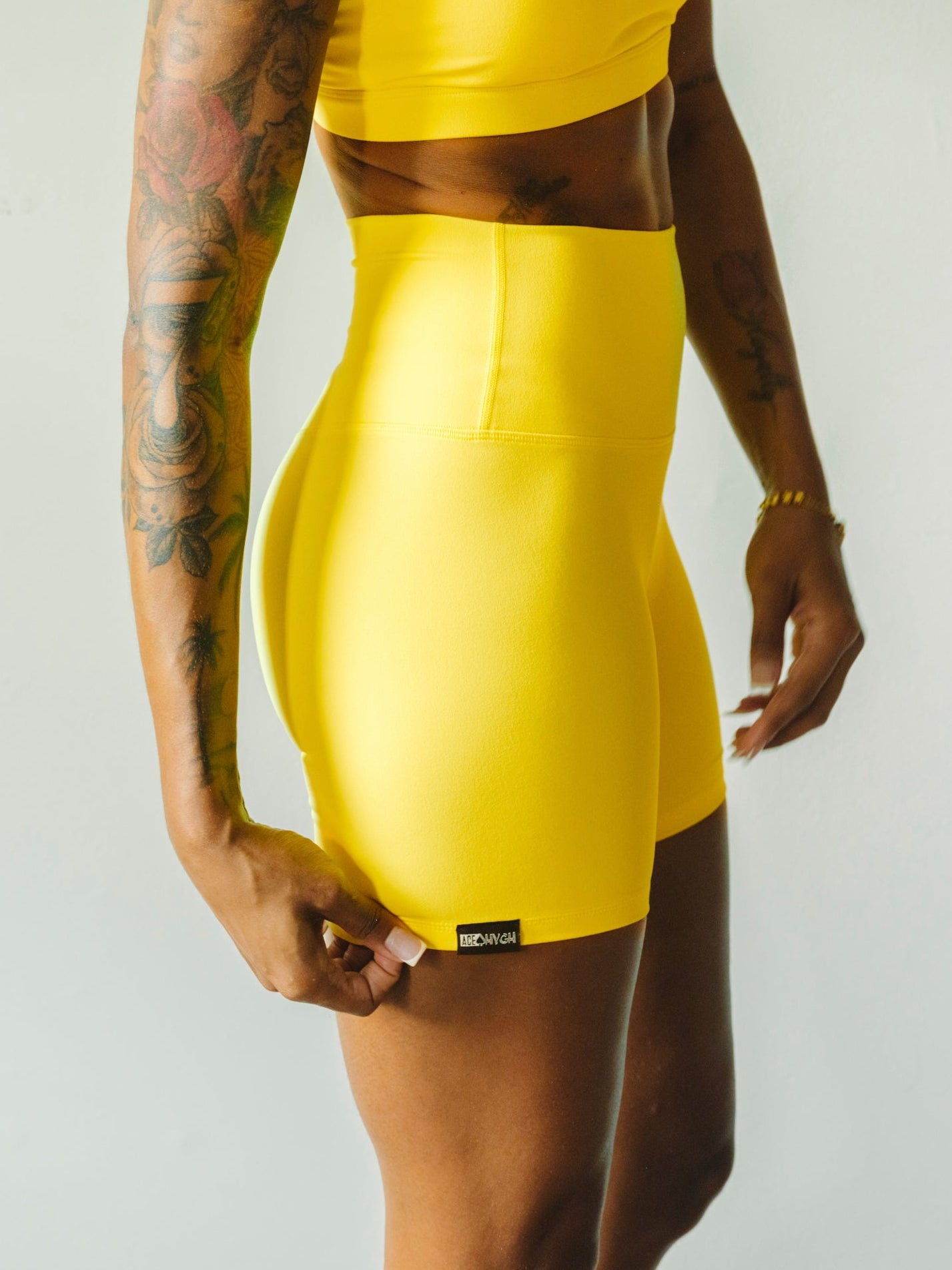 A woman modeling a bright yellow athletic two-piece set consisting of a sports bra and matching high-waisted shorts, set against a plain light gray studio background.