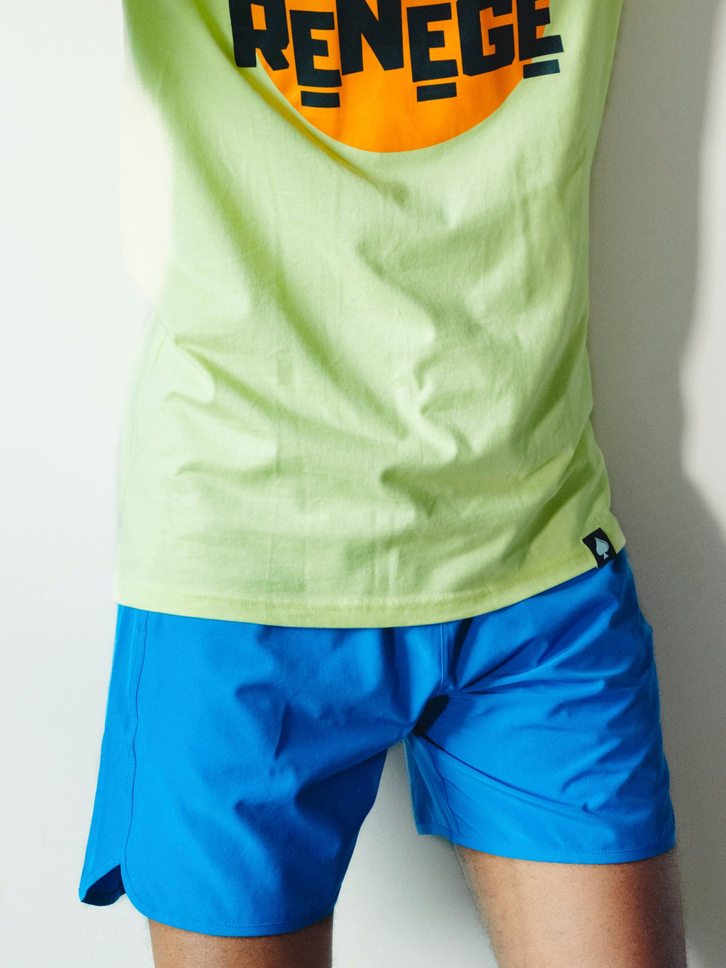 A man wearing a lime green t-shirt with a large orange and black 'DON'T RENEGE' graphic, matching green cap, and blue shorts, posing against a white wall.