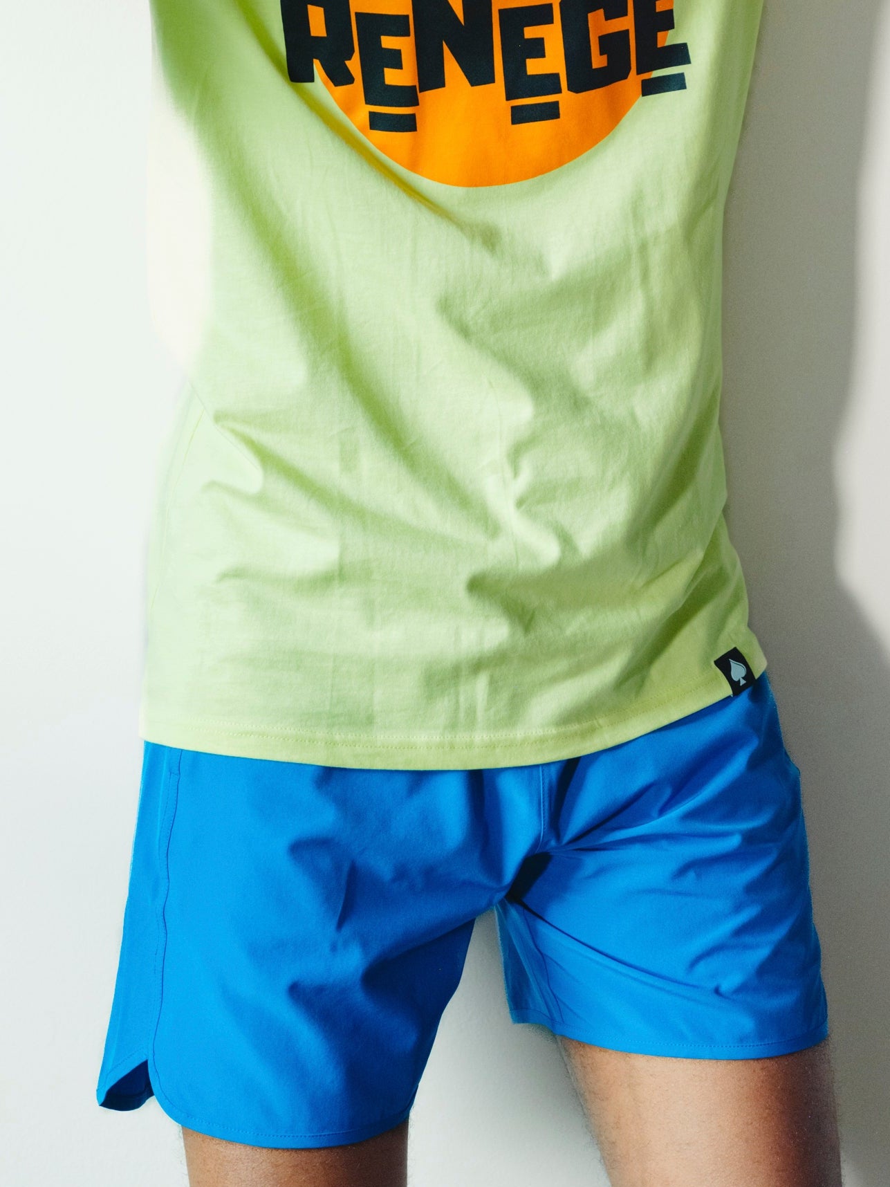 A man wearing a lime green t-shirt with a large orange and black 'DON'T RENEGE' graphic, matching green cap, and blue shorts, posing against a white wall.