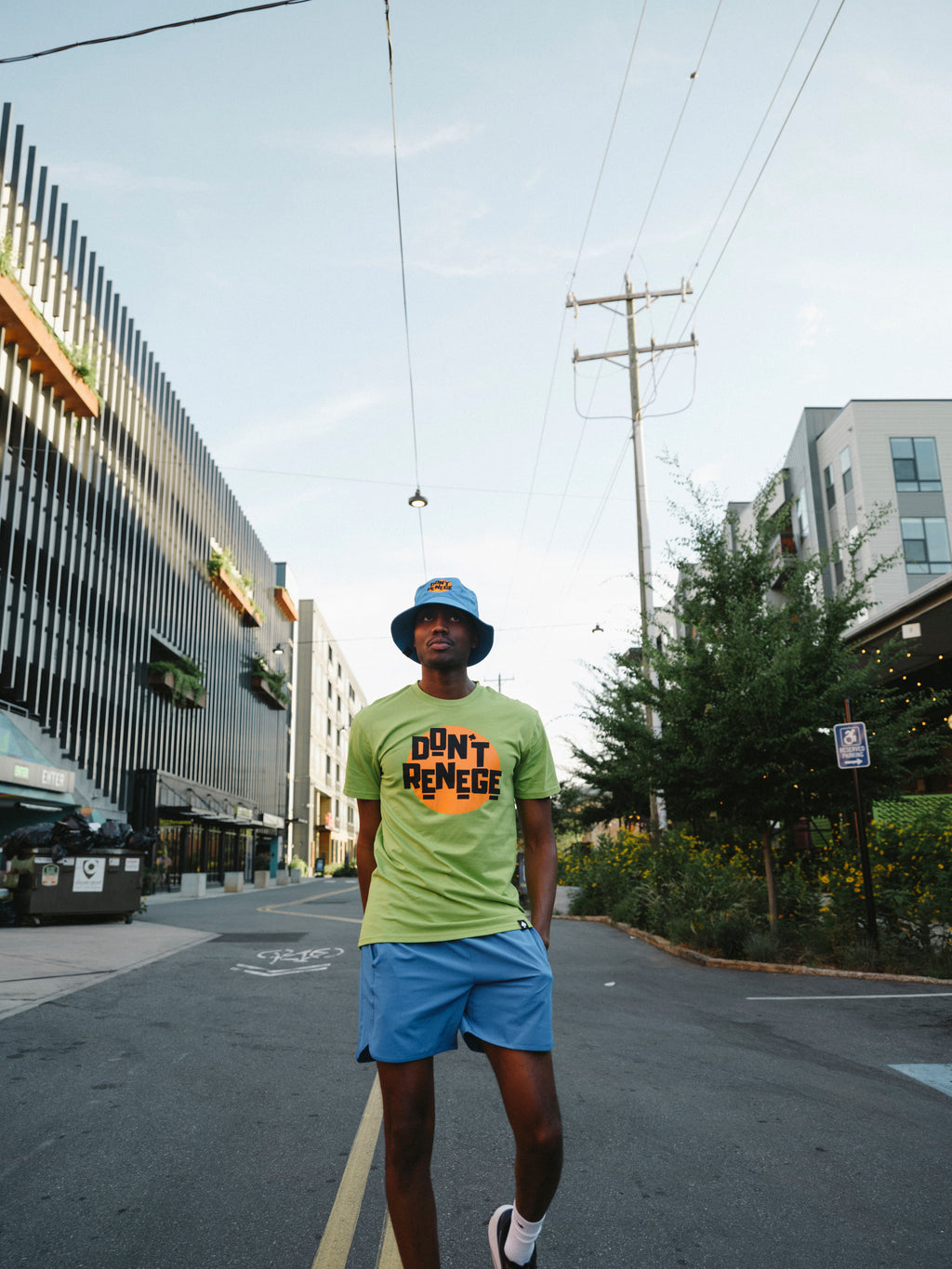 A man walking down an urban street wearing a bright lime green graphic t-shirt with 'DON'T RENGE' printed on the front, blue shorts, and a blue bucket hat.