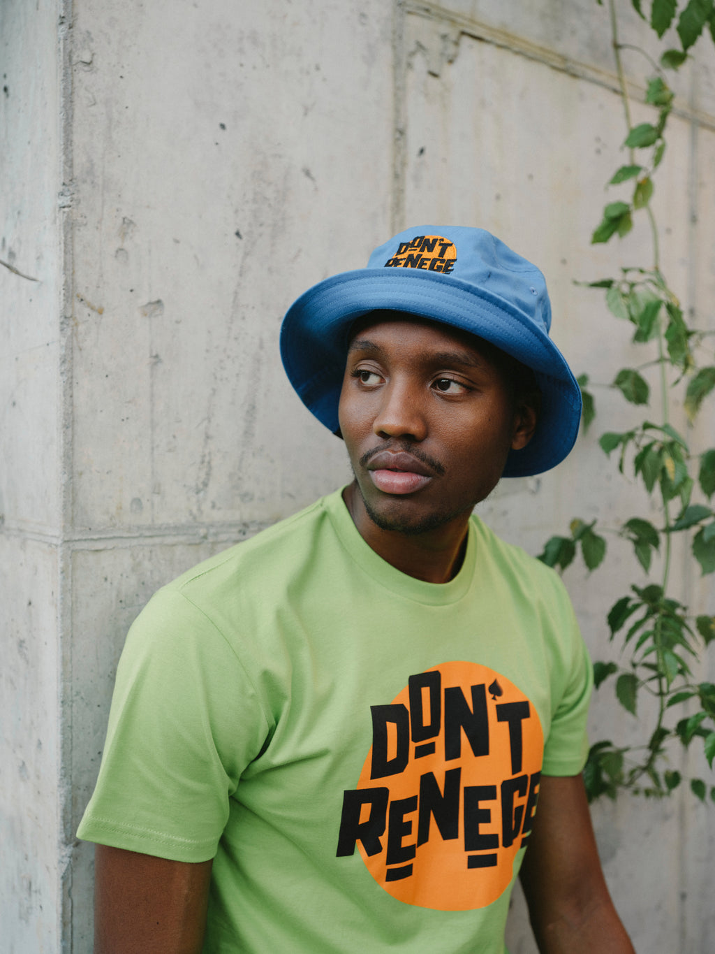 A man wearing a light blue bucket hat and a lime green t-shirt with a bold black and orange graphic that reads 'DON'T RENEGUE', posed against a textured concrete wall with green vines on the right.