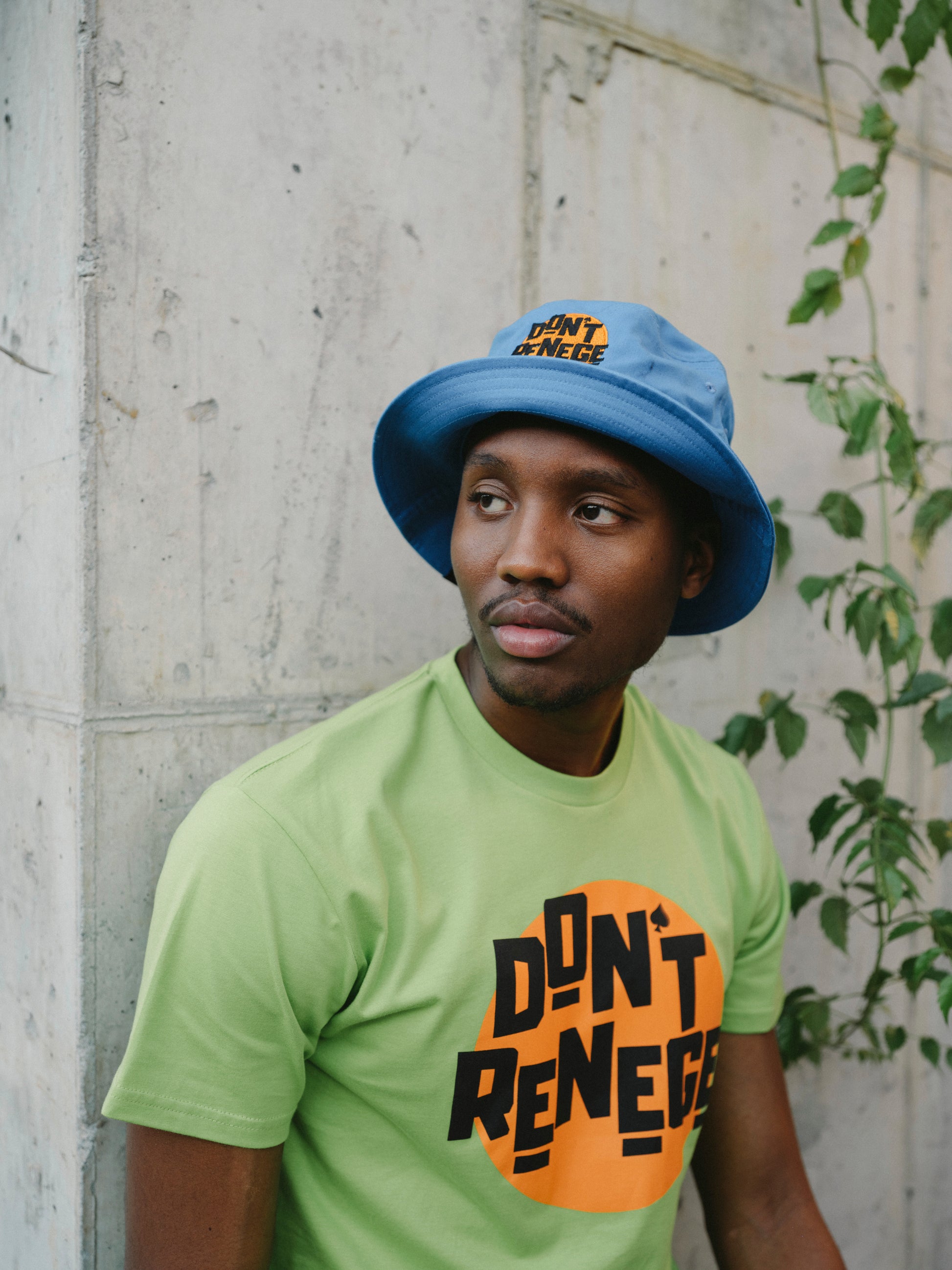 A man wearing a light blue bucket hat and a lime green t-shirt with a bold black and orange graphic that reads 'DON'T RENEGUE', posed against a textured concrete wall with green vines on the right.